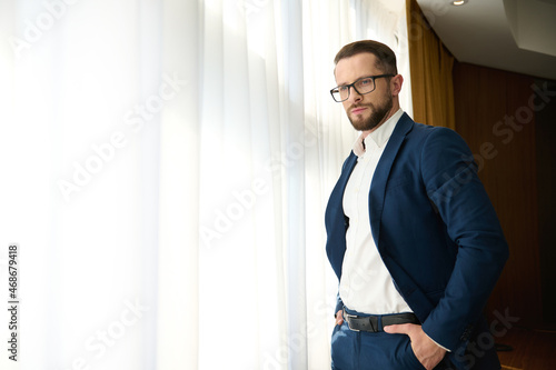 Caucasian successful businessman traveler wearing eyeglasses stands with hands in suit pockets and looking thoughtfully at the cityscape through the hotel balcony window glass during a business trip