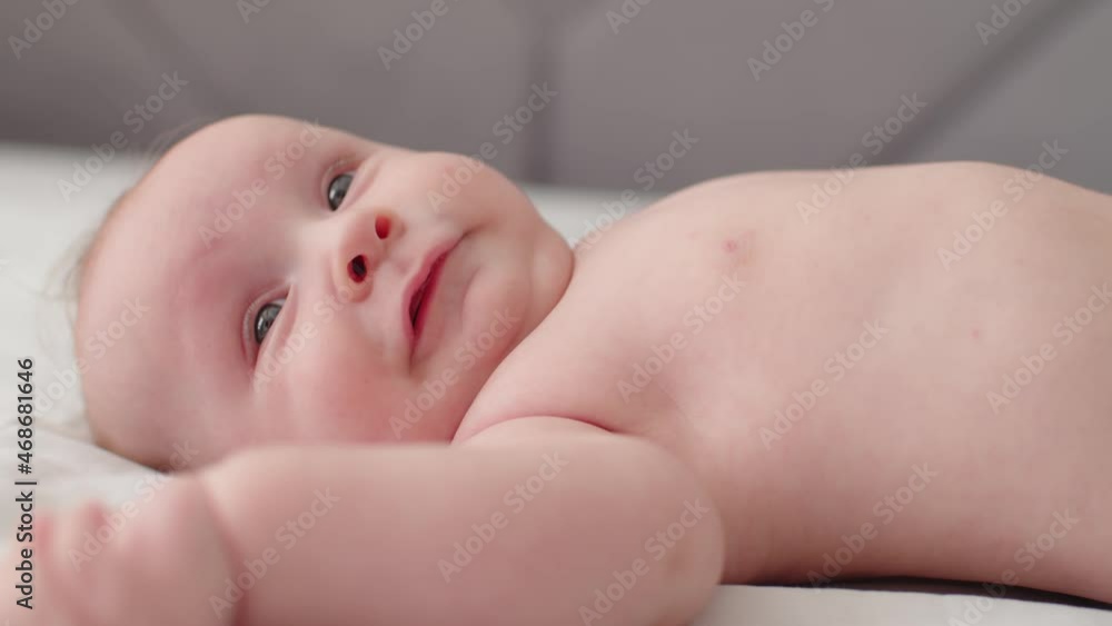 Happy Beautiful newborn baby, closeup face. Cute tiny baby with bare belly looks up, slow motion. Active newborn boy lies on the bed and moves his arms. Low angle view of a chubby baby.. Happy Beautiful newborn baby, closeup face. Cute tiny baby with bare belly looks up, slow motion. Active newborn boy lies on the bed and moves his arms. Low angle view of a chubby baby..