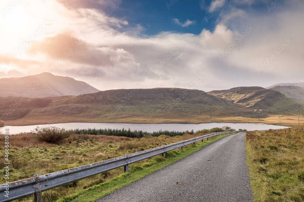 Small narrow asphalt road in beautiful nature setting. Road goes downhill into amazing mountains. Connemara, Ireland. Travel and tourism concept. Metal safety barrier on left side. Dramatic sky