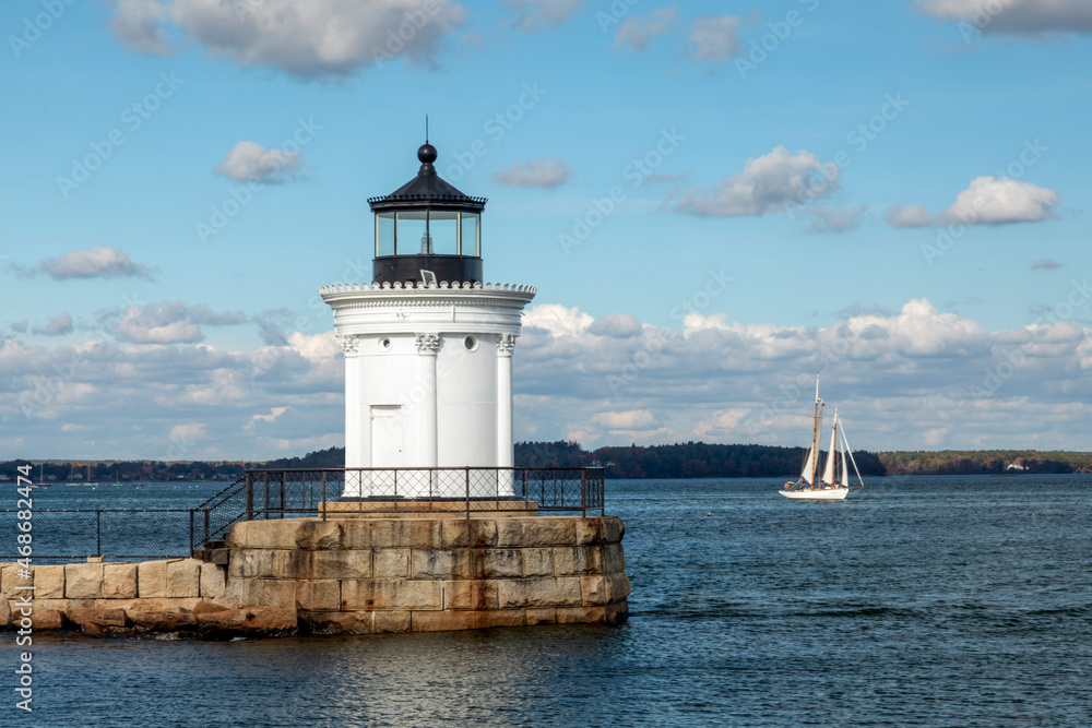 Resembling a Greek monument with Corinthian columns, the Portland Breakwater Lighthouse, also called Bug Light, marks the harbor at Portland, Maine with a sailboat in the distance.