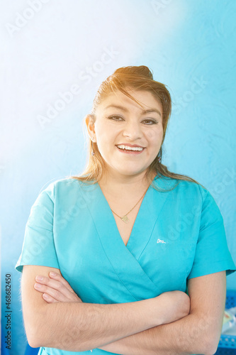 portrait of Dentist latina woman wearing turquoise blue dentist pajamas, on a blue background, with arms crossed.