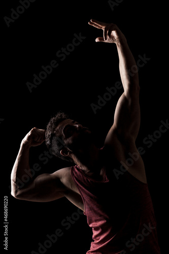 Side lit muscular Caucasian man silhouette. Athlete in red shirt posing against black background.