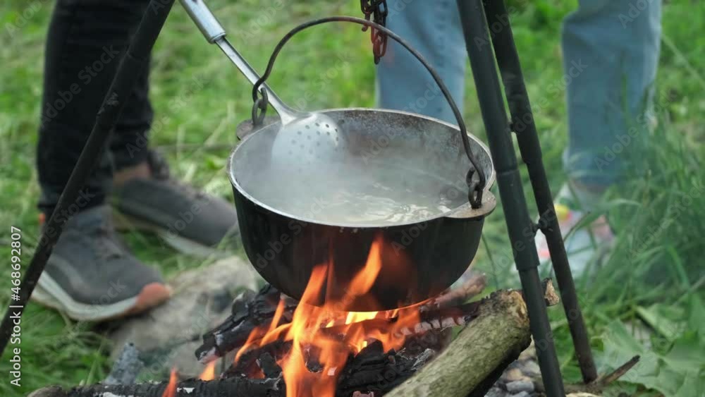 Black large tourist pot hangs over burning flame of campfire in the forest. Cooking dinner on wild hike outdoor. Cauldron of hot food is heated over fire. Outdoor travel. Boil of hot meal in camp. Black large tourist pot hangs over burning flame of campfire in the forest. Cooking dinner on wild hike outdoor. Cauldron of hot food is heated over fire. Outdoor travel. Boil of hot meal in camp.