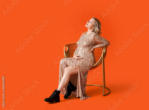 Stylish young pregnant woman sitting in chair on red background