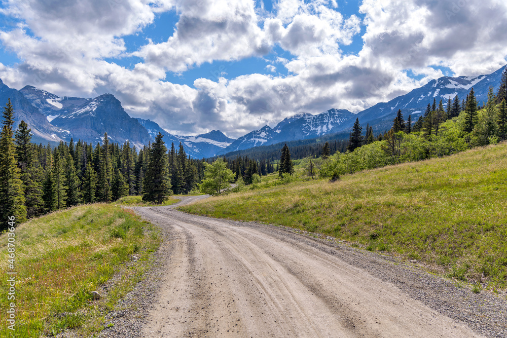Spring Mountain Road - A dirt country road winding in Cut Bank Valley towards high peaks of Lewis Range on a sunny Spring Evening in Glacier National Park, Montana, USA.