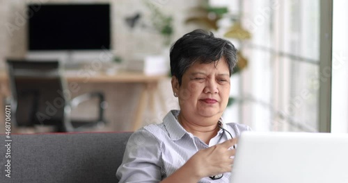 Exhausted mature Asian woman with headache rubbing eyes and touching head while sitting on sofa and working on remote project on laptop at home