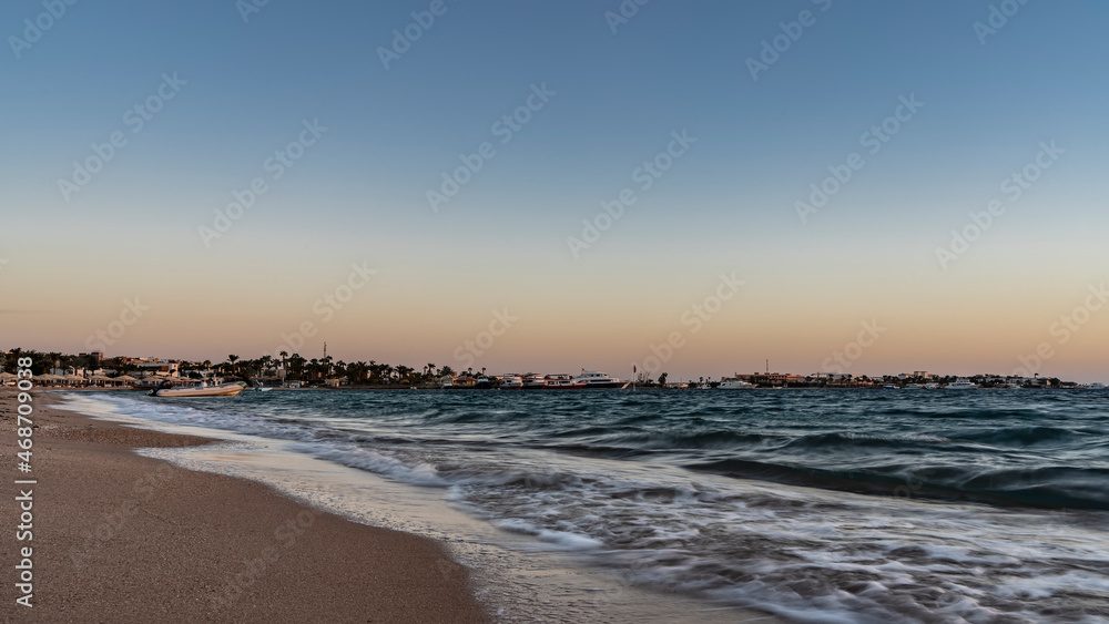 Early morning on the Red Sea beach. Turquoise waves foam on the sandy beach. In the distance you can see sun umbrellas, palm trees, boats. The sky is highlighted in orange. Egypt