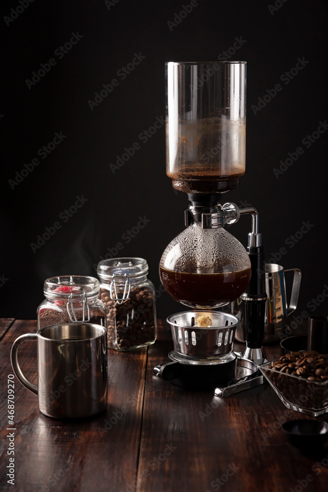 Vacuum coffee maker also known as vac pot, siphon or syphon coffee maker. Metallic cup and toasted coffee beans on rustic wooden table Vacuum coffee maker also known as vac pot, siphon or syphon coffee maker. Metallic cup and toasted coffee beans on rustic wooden table
