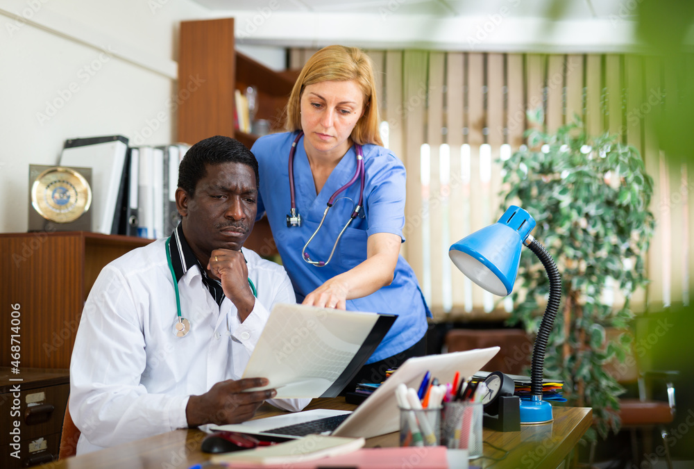 Obraz premium Two focused professional doctors working at table in office of clinic, discussing medical histories of patients