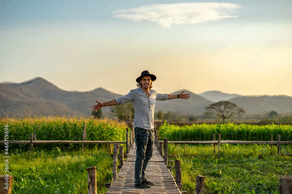 Happy young handsome man and a hat while traveling with glad positive expression in Asia. Attractive male in harmony with the field nature raises his hands to sunset or sunrise and mountain landscape