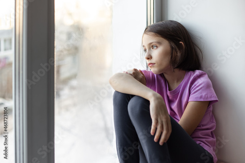 A sad, serious child sits on the windowsill and sadly looks out the window. Waiting, sadness, thoughtfulness.