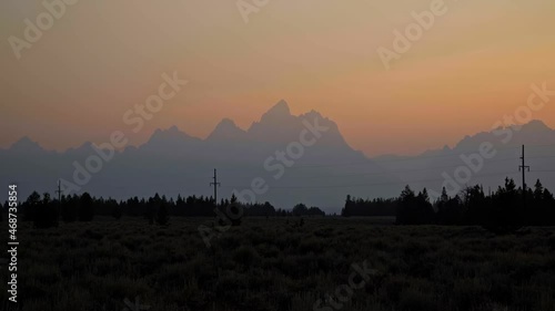 Stunning landscape nature tilting down shot of the Grant Teton mountain range during a hazy summer sunset with pine trees and power lines below near Jackson Hole, Wyoming, USA. photo