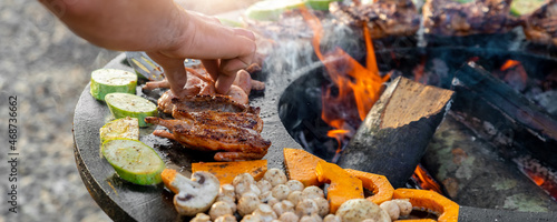 Close-up detail view of chef flipping by tongs tasty crispy marinated quails and vegetables grilled at round steel iron firepit hearth table surface brazier with burning firewoods. Barbecue yard home