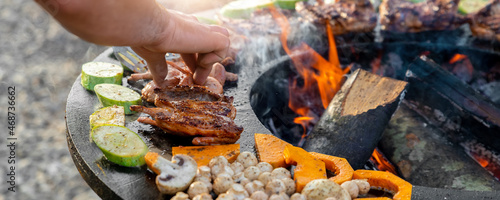 Close-up detail view of chef flipping by tongs tasty crispy marinated quails and vegetables grilled at round steel iron firepit hearth table surface brazier with burning firewoods. Barbecue yard home photo