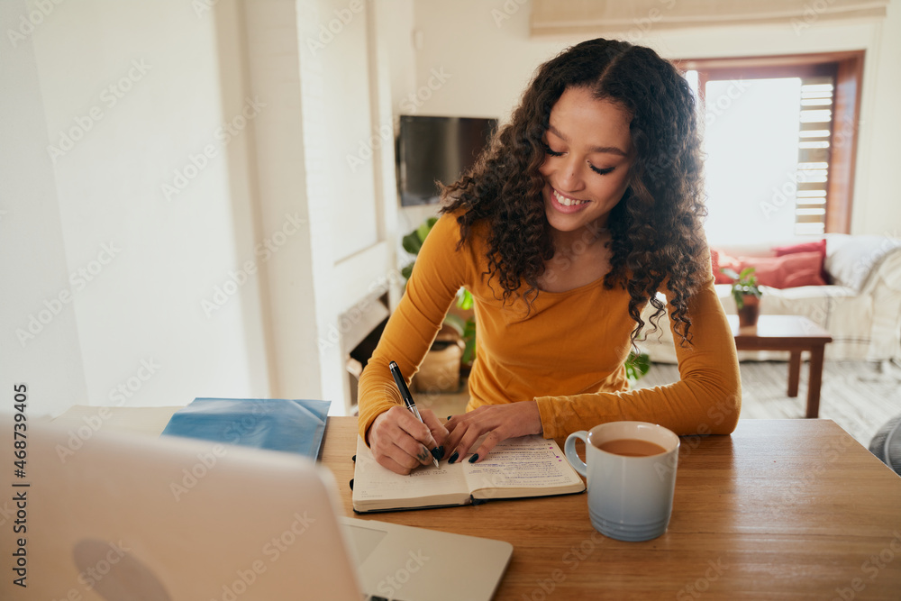 Multi-cultural female happily writing in notebook. Young professional remote working with laptop in modern apartment Multi-cultural female happily writing in notebook. Young professional remote working with laptop in modern apartment