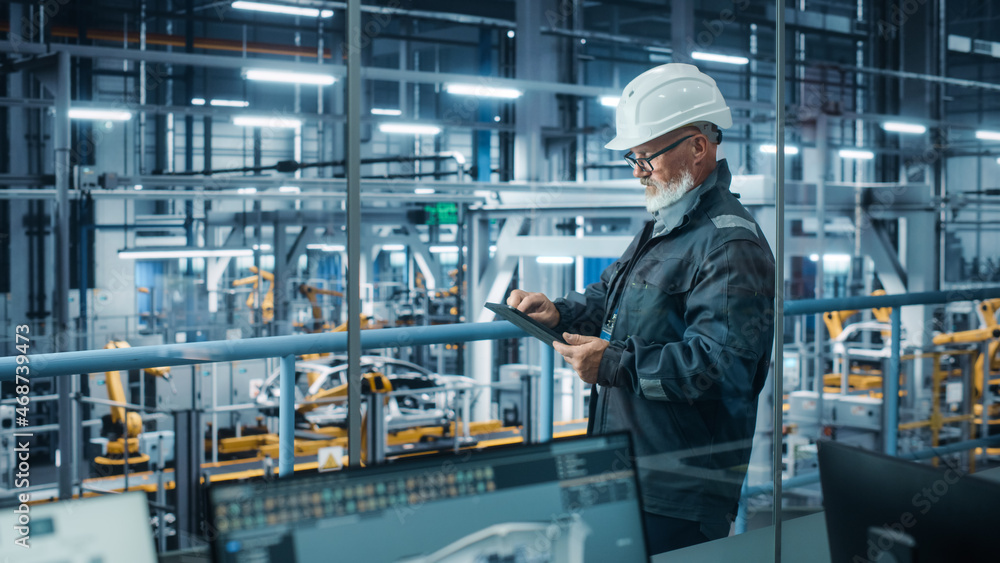 Car Factory: Male Automotive Engineer Wearing Hard Hat, Standing Using Tablet Computer. Monitoring, Control, Equipment Production. Automated Robot Arm Assembly Line Manufacturing Electric Vehicles