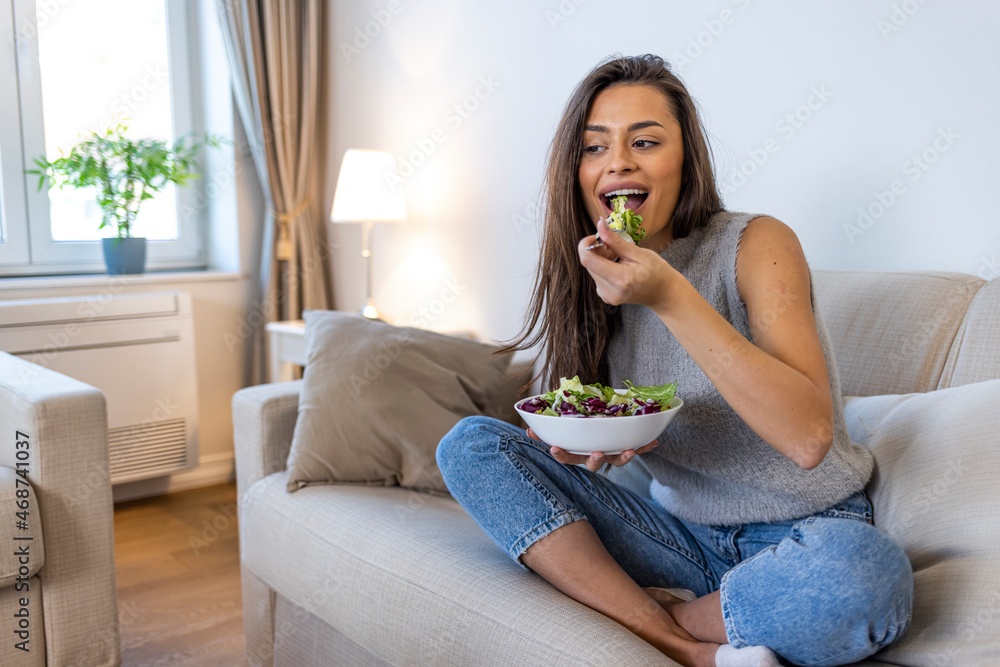 Photo of young woman enjoying a delicious salad while standing in her living room at home during the day. Young and happy woman eating healthy salad with green fresh ingredients indoors Photo of young woman enjoying a delicious salad while standing in her living room at home during the day. Young and happy woman eating healthy salad with green fresh ingredients indoors