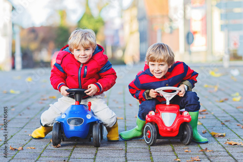 Two little kids boys in colorful clothes and rain boots driving toy cars. Twins making competition, outdoors. Active leisure for children on autumn day.