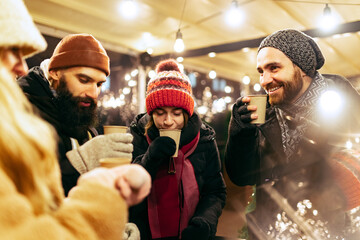 Close-up happy smiling friends with cups of mulled wine spending time together at winter fair at evening. Holidays, Christmas concept