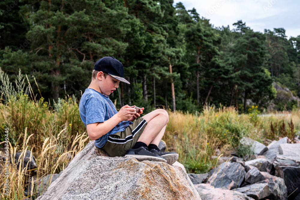 Side view of young boy sitting on rock at seashore looking at mobile phone. Forest and rocks in the background. Side view of young boy sitting on rock at seashore looking at mobile phone. Forest and rocks in the background.