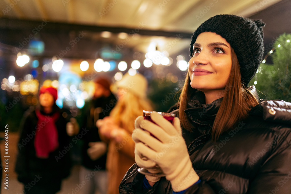 Close-up of young beautiful girl in winter attire drinking hot drinks and dreaming. Spending time with friends at winter fair at evening.