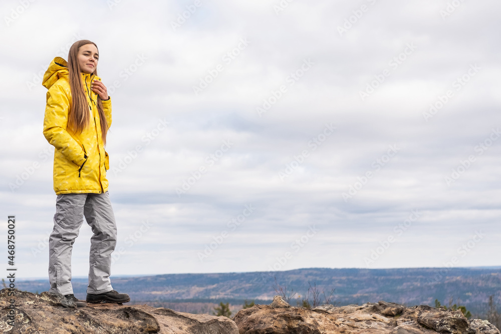 A smiling tourist, in a yellow jacket and gray pants, stands on the top of a mountain in a valley of mountains and admires the beauty of nature.