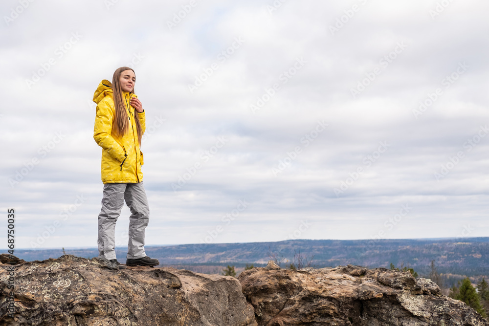 A smiling tourist, in a yellow jacket and gray pants, stands on the top of a mountain in a valley of mountains and admires the beauty of nature.