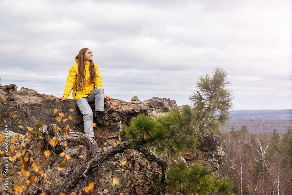 Fototapeta premium A smiling tourist, in a yellow jacket and gray pants, sits on the top of a mountain, in a beautiful valley with mountains and cloudy skies, admiring the beauty of nature.