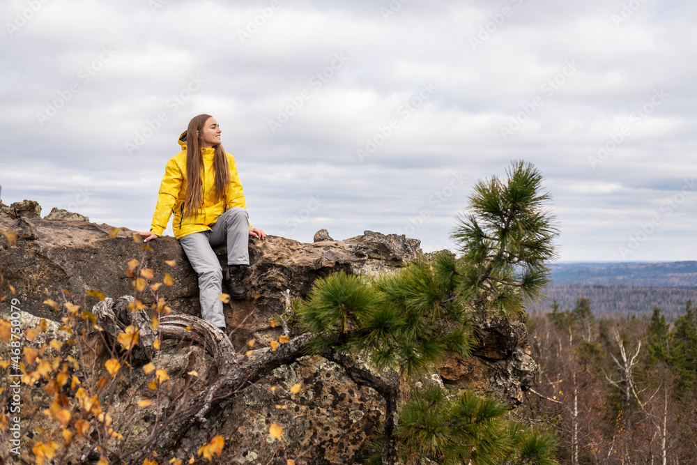 A smiling tourist, in a yellow jacket and gray pants, sits on the top of a mountain, in a beautiful valley with mountains and cloudy skies, admiring the beauty of nature.