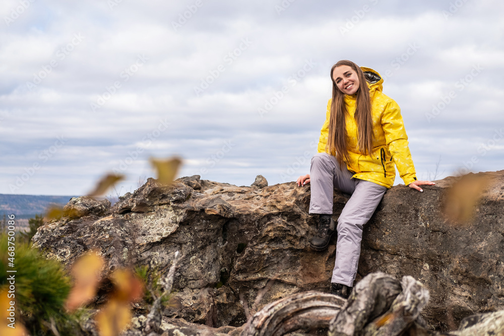 Naklejka premium Smiling tourist, in a yellow jacket and gray pants, sits on the top of a mountain, in a beautiful valley with mountains and a cloudy sky, and looks at the camera.