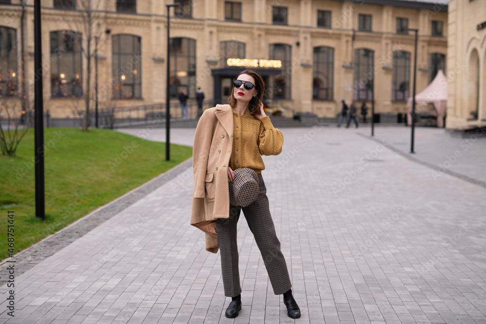 Charming model girl in an autumn beige coat and sunglasses, plaid panama hat standing with face turned holding coat on her shoulders. Retro toned photo