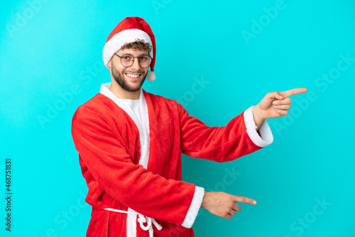 Young man disguised as Santa Claus isolated on blue background pointing finger to the side and presenting a product