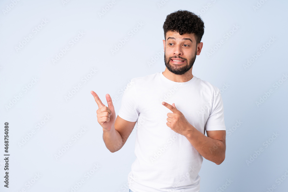 Young Moroccan man isolated on blue background frightened and pointing to the side Young Moroccan man isolated on blue background frightened and pointing to the side