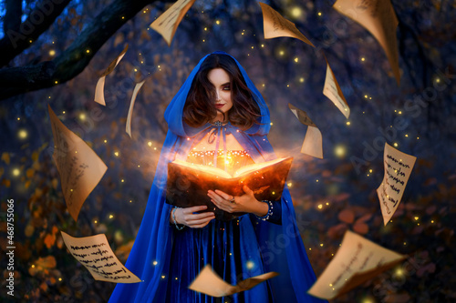Quadro su tela Fantasy woman witch magician in hood holds in hands magic book, bright orange light spells, wind scatters fall sheets paper page levitation