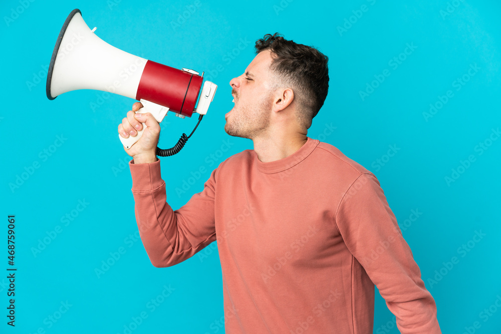 Young caucasian handsome man isolated on blue background shouting through a megaphone to announce something in lateral position Young caucasian handsome man isolated on blue background shouting through a megaphone to announce something in lateral position