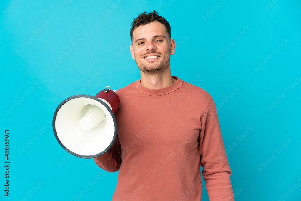 Young caucasian handsome man isolated on blue background holding a megaphone and smiling a lot Young caucasian handsome man isolated on blue background holding a megaphone and smiling a lot