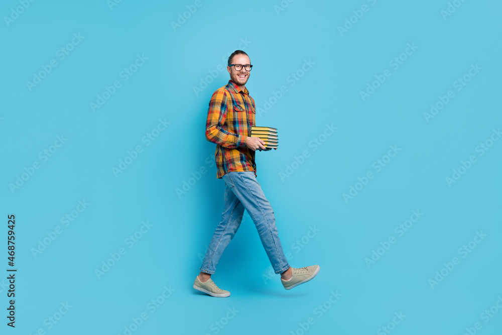 Full length profile photo of funny millennial brunette guy hold book go wear shirt jeans shoes isolated on blue color background Full length profile photo of funny millennial brunette guy hold book go wear shirt jeans shoes isolated on blue color background