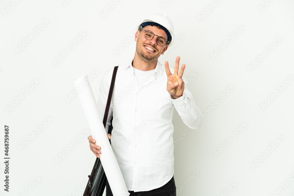 Young architect man with helmet and holding blueprints isolated on white background happy and counting three with fingers Young architect man with helmet and holding blueprints isolated on white background happy and counting three with fingers