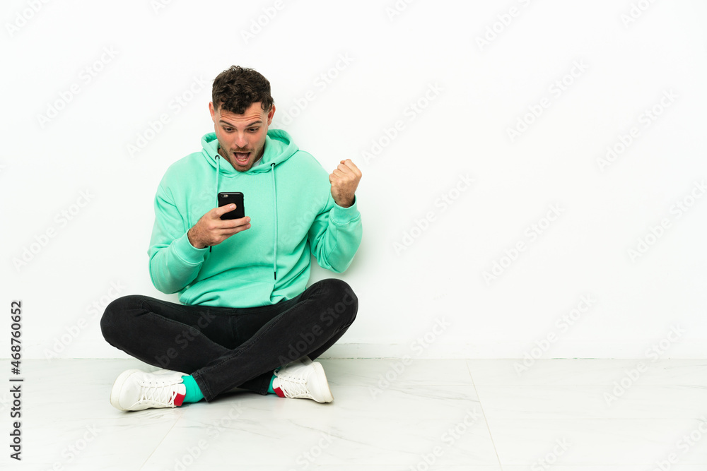 Young handsome caucasian man sitting on the floor surprised and sending a message Young handsome caucasian man sitting on the floor surprised and sending a message