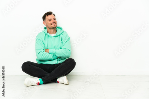 Young handsome caucasian man sitting on the floor with arms crossed and happy