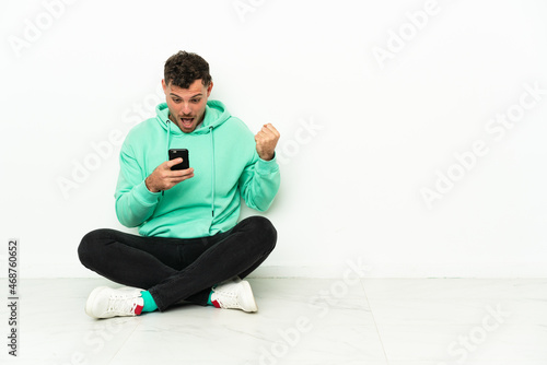 Young handsome caucasian man sitting on the floor surprised and sending a message