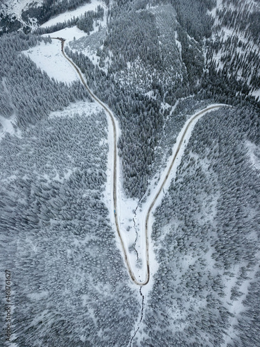 Aerial panorama of a sinuous gravel road winding through the snowed forests of Parang Mountains. Wild coniferous woods are growing on the mountain sides. Carpathia, Romania.
