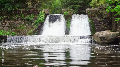 Two waterfalls running over concrete slope and splashing in lake forming white foam. Wind forming ripples on the lake surface. Surrounded by gray stones and green trees