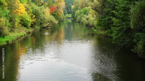 Autumn landscape of flowing river surrounded by green, yellow and orange trees. Tree reflections in the water and forest in the background. Wind blowing across the water surface and tree leaves