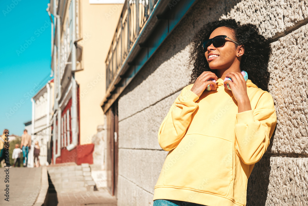 Beautiful black woman with afro curls hairstyle.Smiling model in yellow hoodie.Sexy carefree female enjoying listening music in wireless headphones.Posing on street background near wall in sunglasses Beautiful black woman with afro curls hairstyle.Smiling model in yellow hoodie.Sexy carefree female enjoying listening music in wireless headphones.Posing on street background near wall in sunglasses