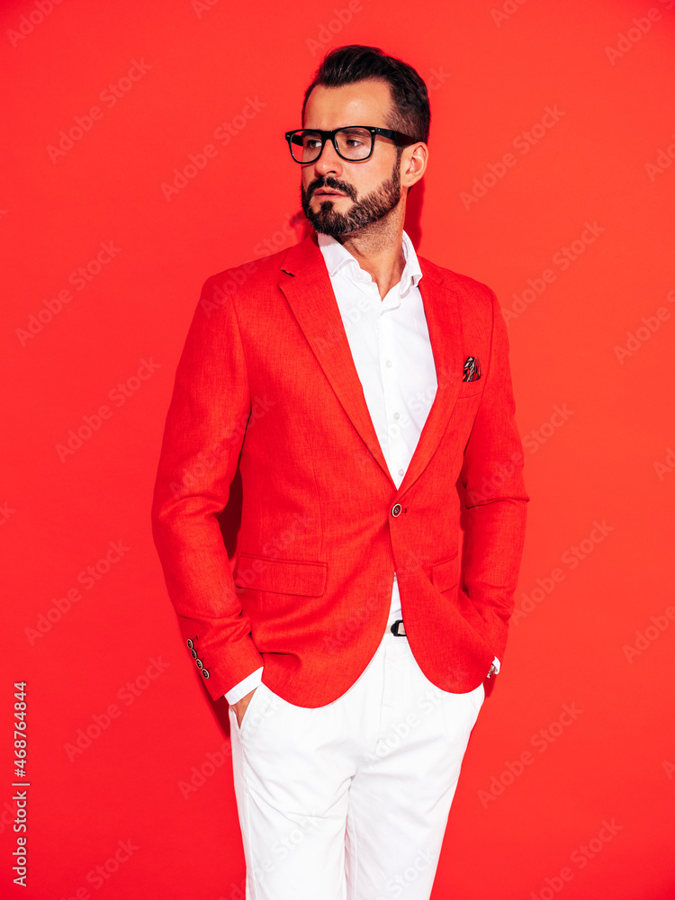 Portrait of handsome confident stylish hipster lambersexual model. Sexy modern man dressed in elegant suit. Fashion male posing in studio near red wall in spectacles
