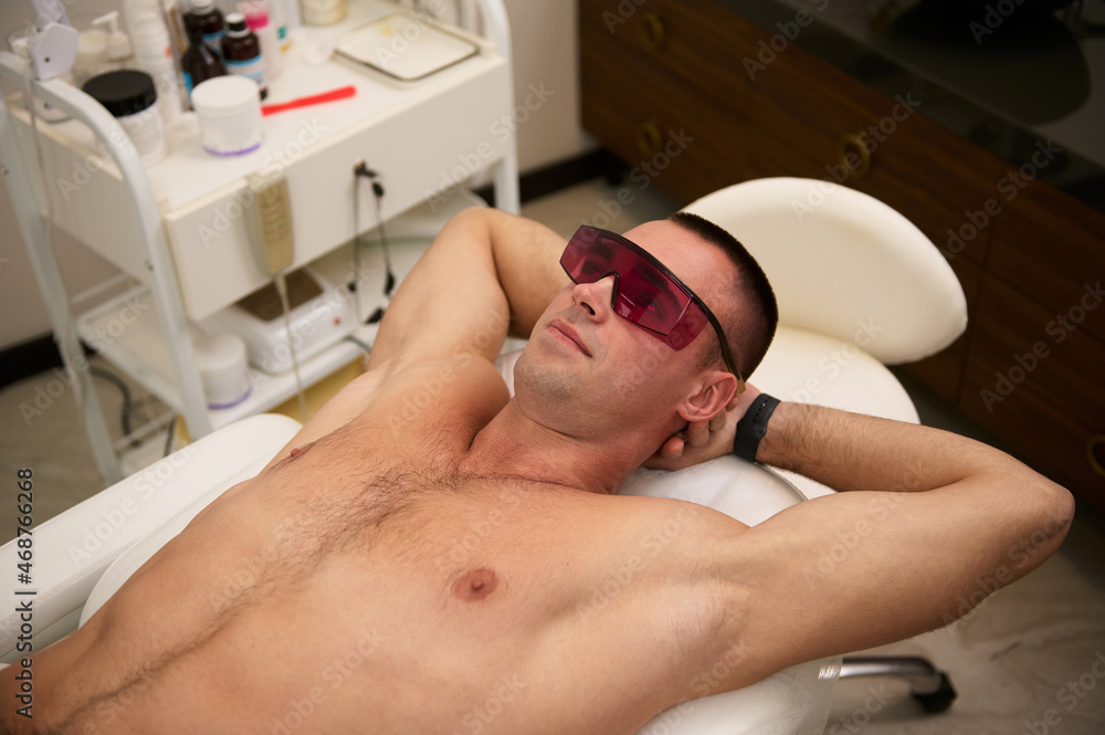Young attractive manly sporty man wearing UV protective goggles, lying on a massage table of spa salon during laser hair removal procedure. Masculine beauty spa treatment concept. Male waxing Young attractive manly sporty man wearing UV protective goggles, lying on a massage table of spa salon during laser hair removal procedure. Masculine beauty spa treatment concept. Male waxing