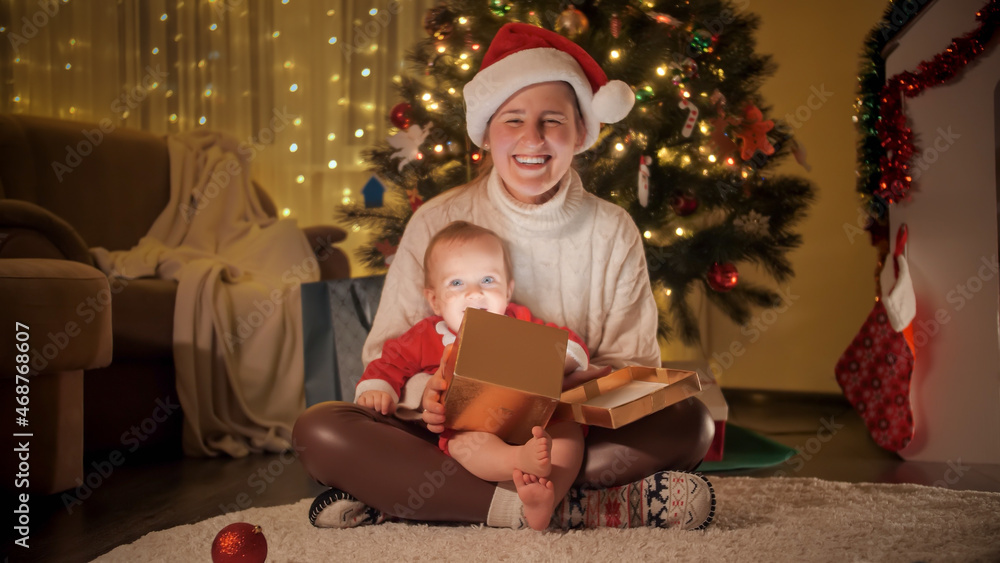 Happy smiling mother holding Christmas gift and baby son while sitting on carpet under Christmas tree