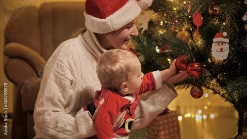 Little baby boy with mother in Santa Claus hat decorating Christmas tree with baubles. Families and children celebrating winter holidays.