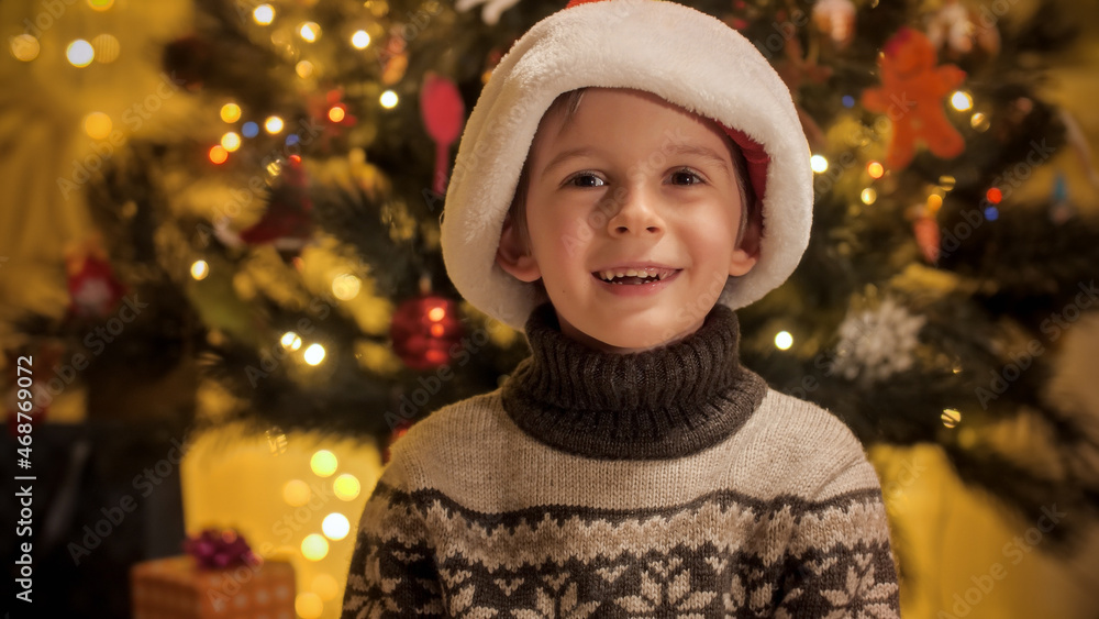 Cute smiling boy in knitted warm sweater and Santa hat celebrating New Year next to Christmas tree Cute smiling boy in knitted warm sweater and Santa hat celebrating New Year next to Christmas tree